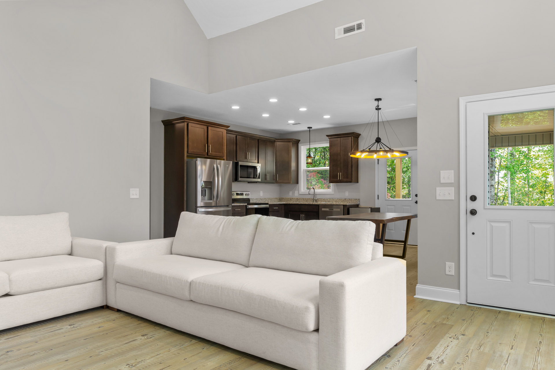 White couch with pillows in a living room featuring hardwood floors, a ceiling chandelier, and large windows showing trees outside; white door with glass panel and built-in