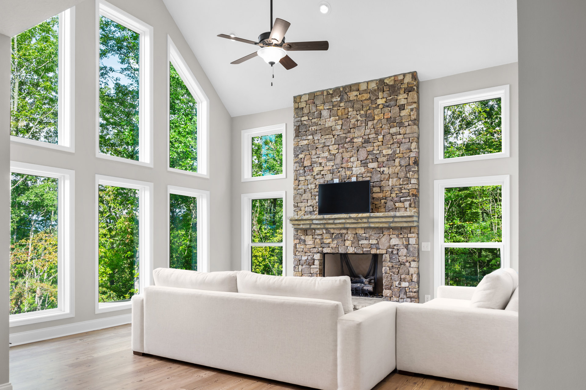 Living room with white couches, stone fireplace, wall-mounted TV, ceiling fan with light, large window showing trees outside