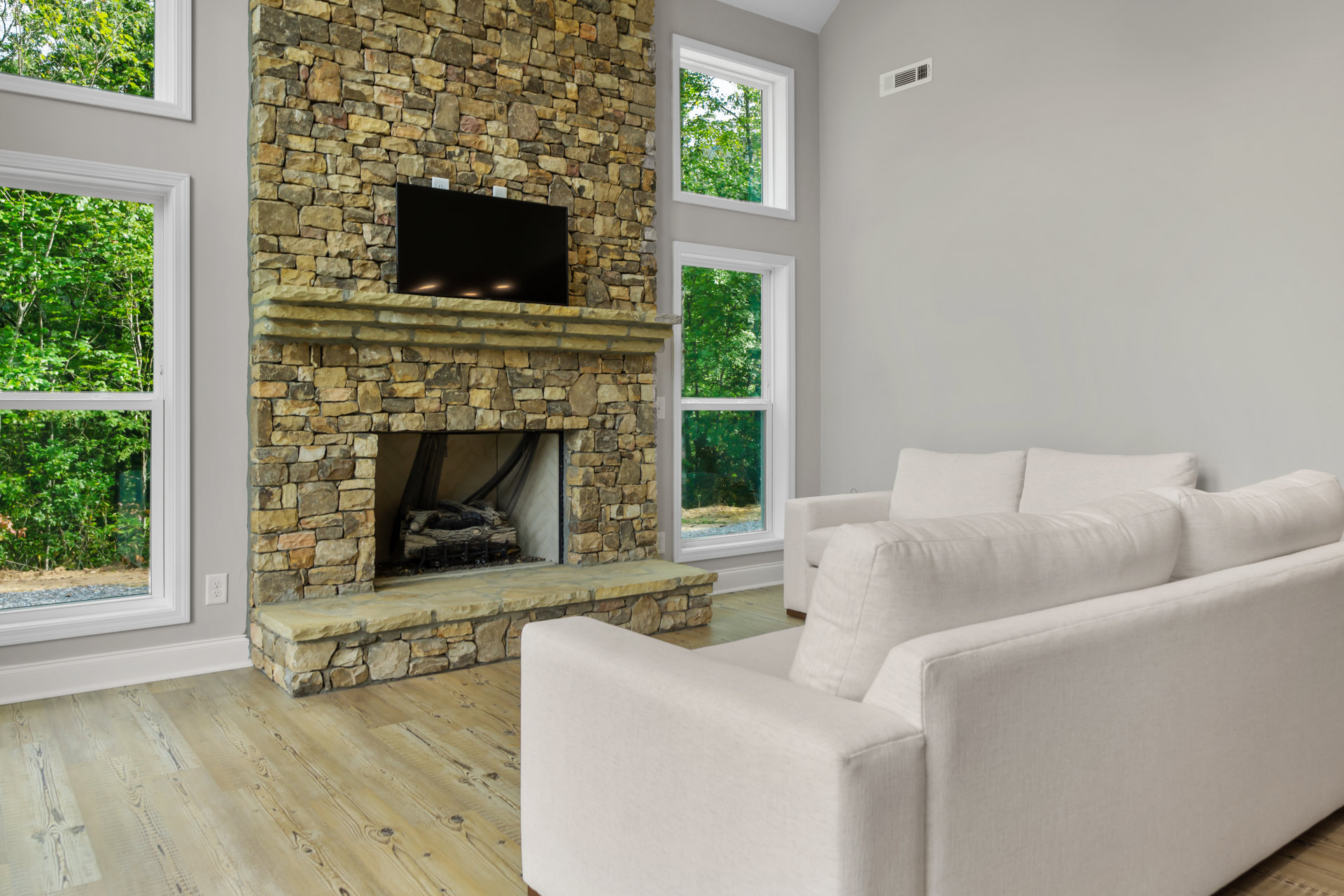Living room with stone fireplace, mounted TV, white couches, large window showing trees, and wood flooring