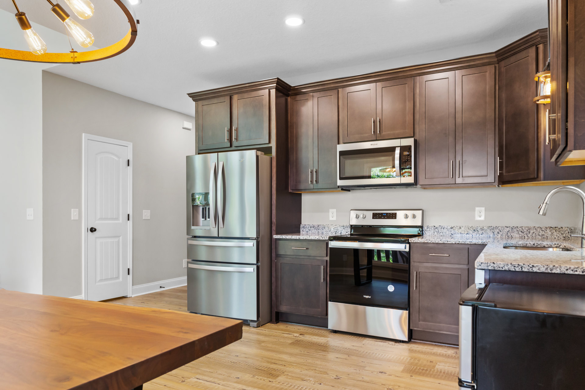 Kitchen with wooden dining table, stainless steel double-door refrigerator, black and silver oven, microwave with illuminated window, light bulb fixture, wood cabinetry, and stone