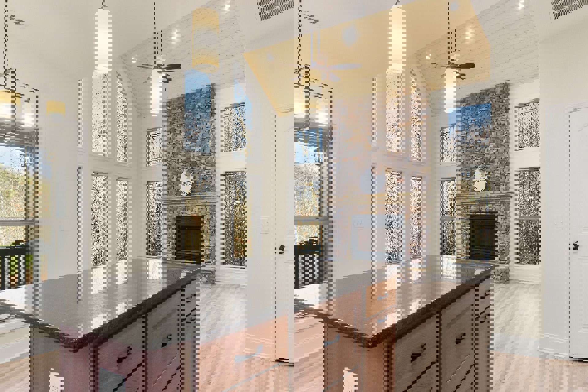 Kitchen with stone fireplace featuring glass door, ceiling fan, wood cabinetry and drawers, countertop, window overlooking trees, decorative sign with bow, and a white rectangular