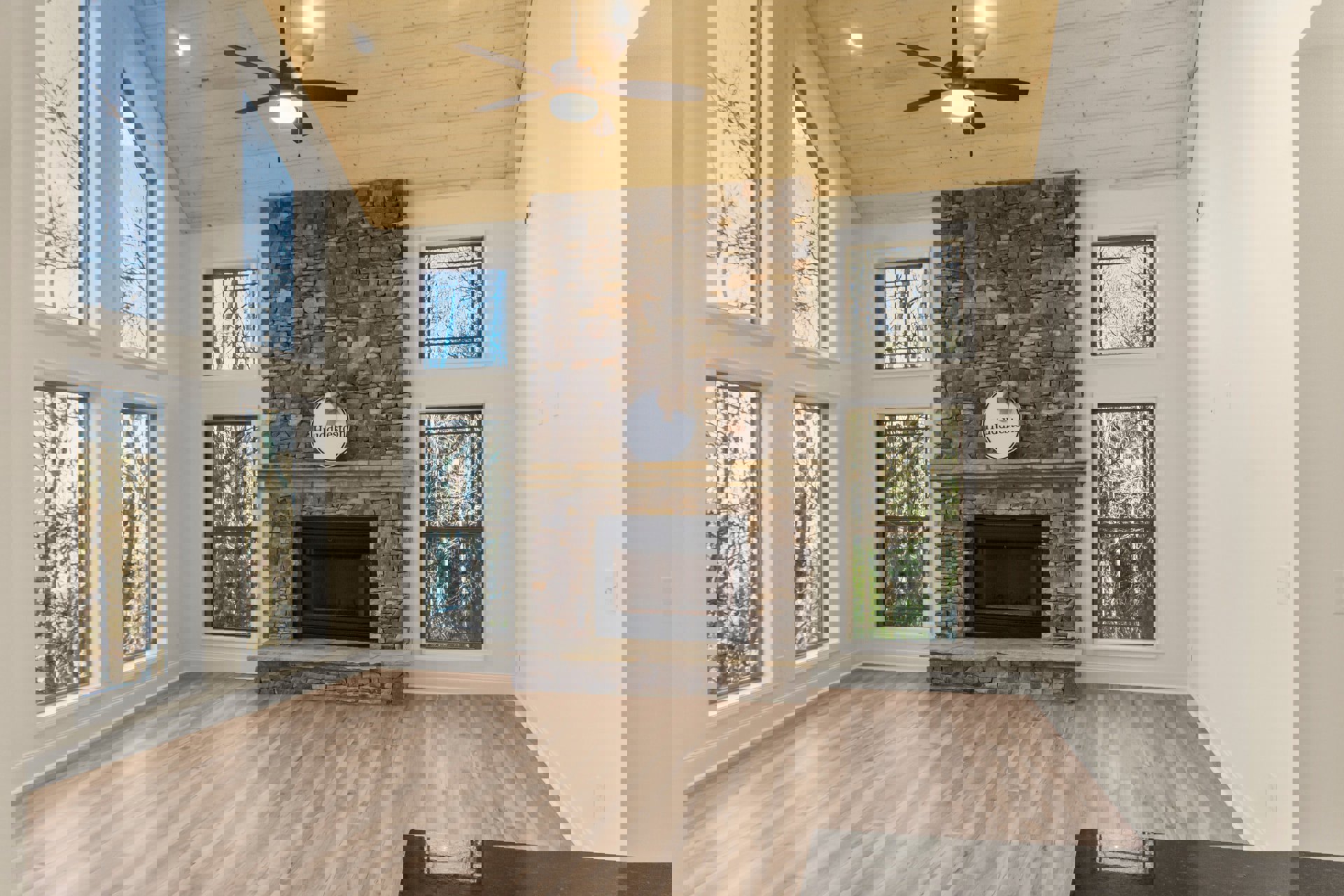 Living room with stone fireplace, wood flooring, ceiling fan with light, large window showing trees, and decorative sign with bow on mantel.