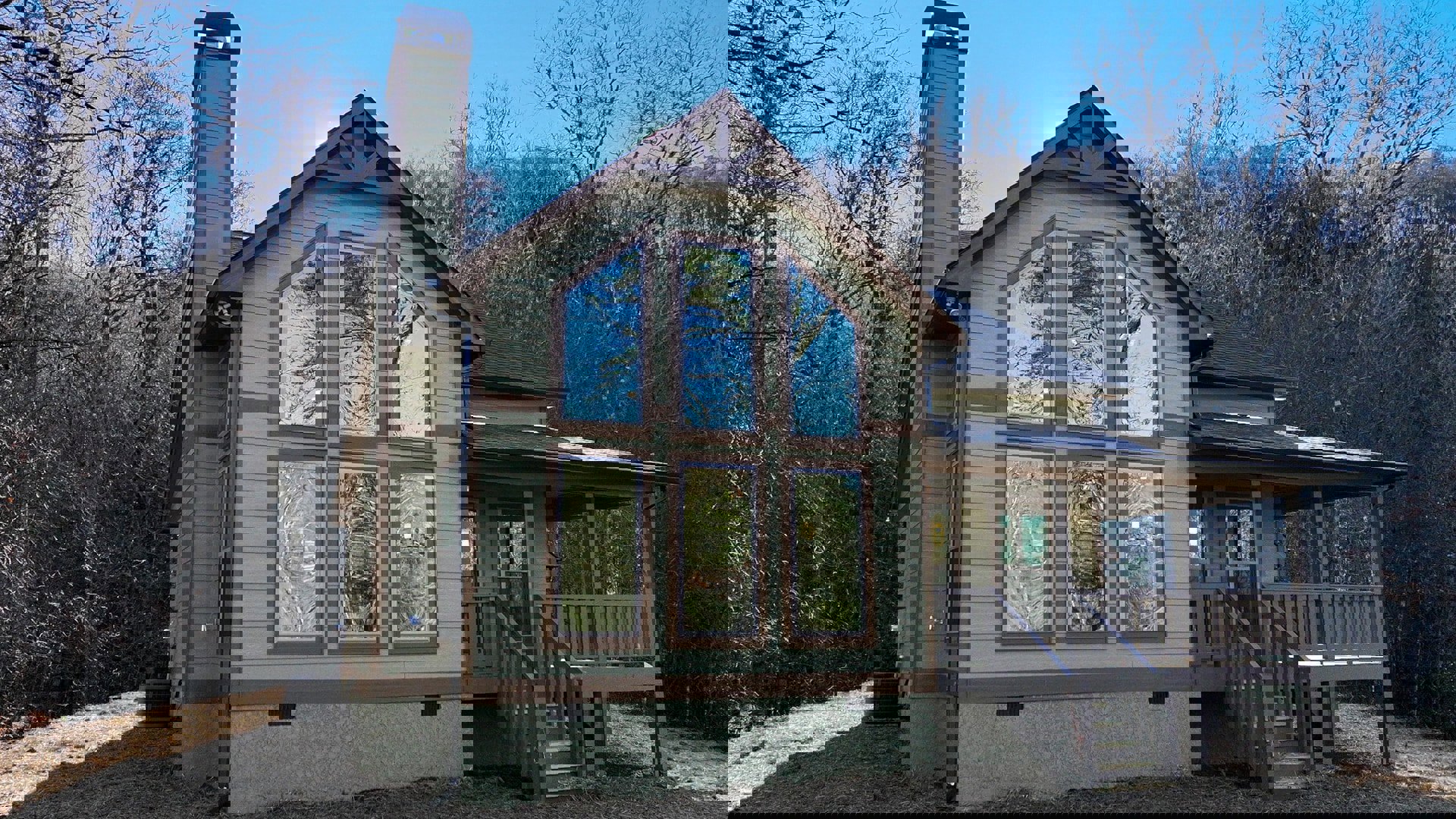 Two-story home with multiple large windows reflecting trees, prominent brick chimney, covered porch with white railing, and green and brown window frames
