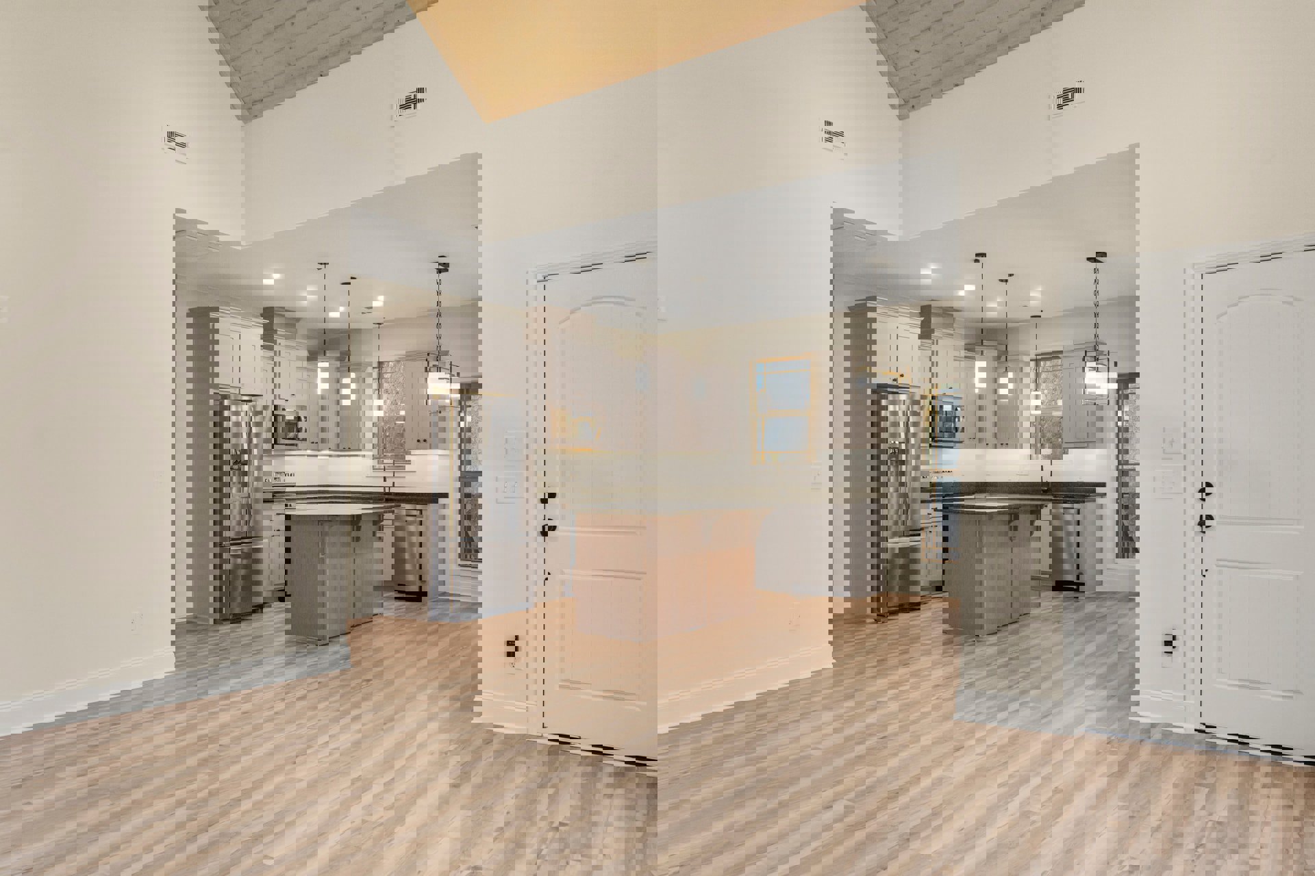 Open kitchen and dining area featuring wood flooring, stainless steel double-door refrigerator, white door with black knobs, wooden cabinetry with black countertops, wall vent, and