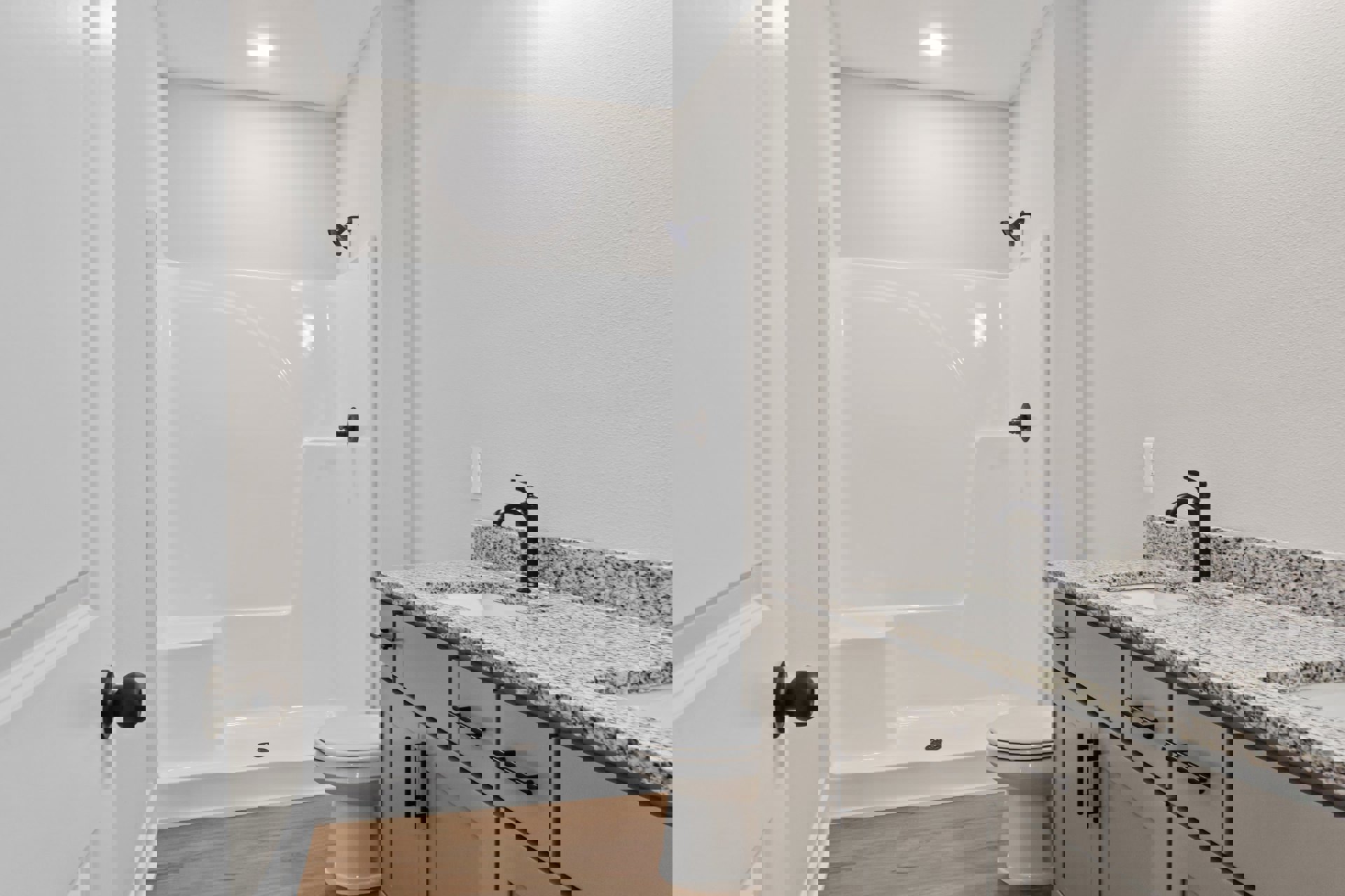 White tile shower with glass door open, white toilet with closed seat, white vanity cabinets with black handles, undermount sink, black door knob on white wall, chrome shower head.