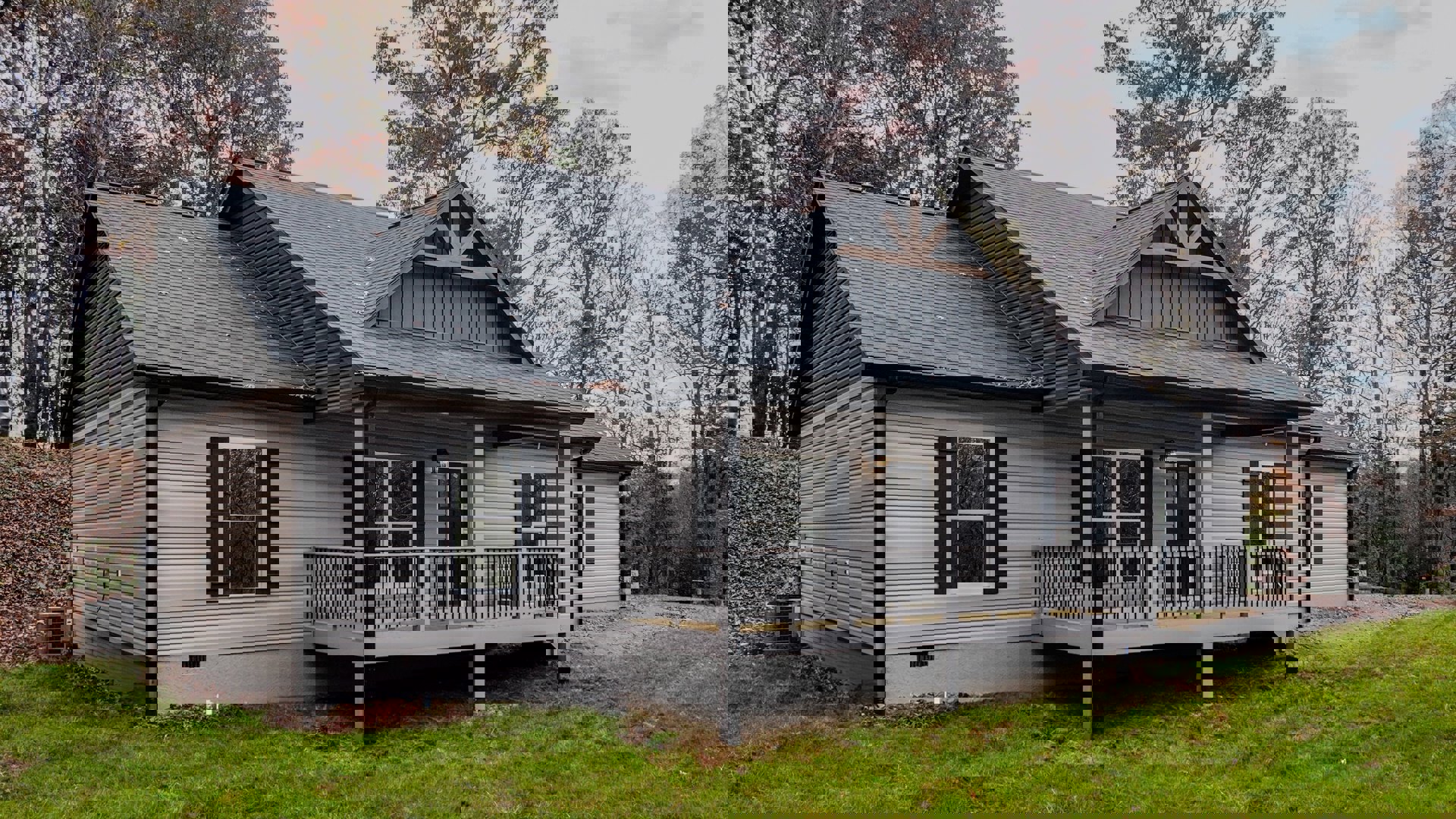 Two-story home with gray siding, white-framed windows, covered front porch, and elevated deck featuring metal railing, surrounded by grass and trees under a clear sky