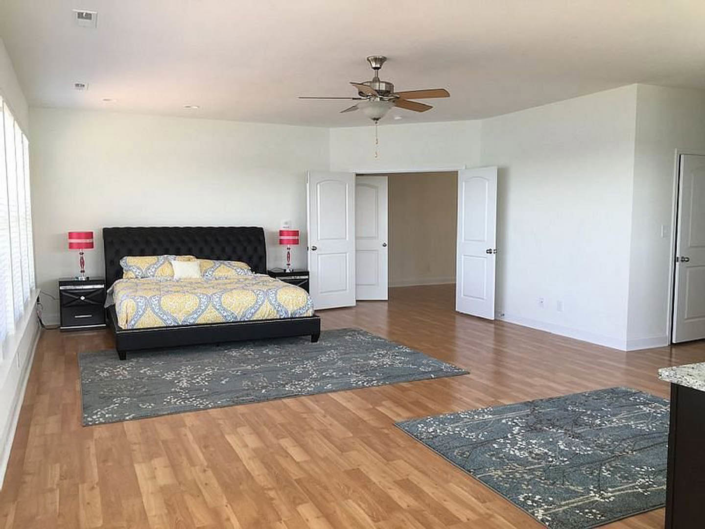 Bedroom with a large bed featuring a black headboard, patterned rug on wood laminate flooring, ceiling fan with light fixture, white pillow on a couch, and close-up of a modern