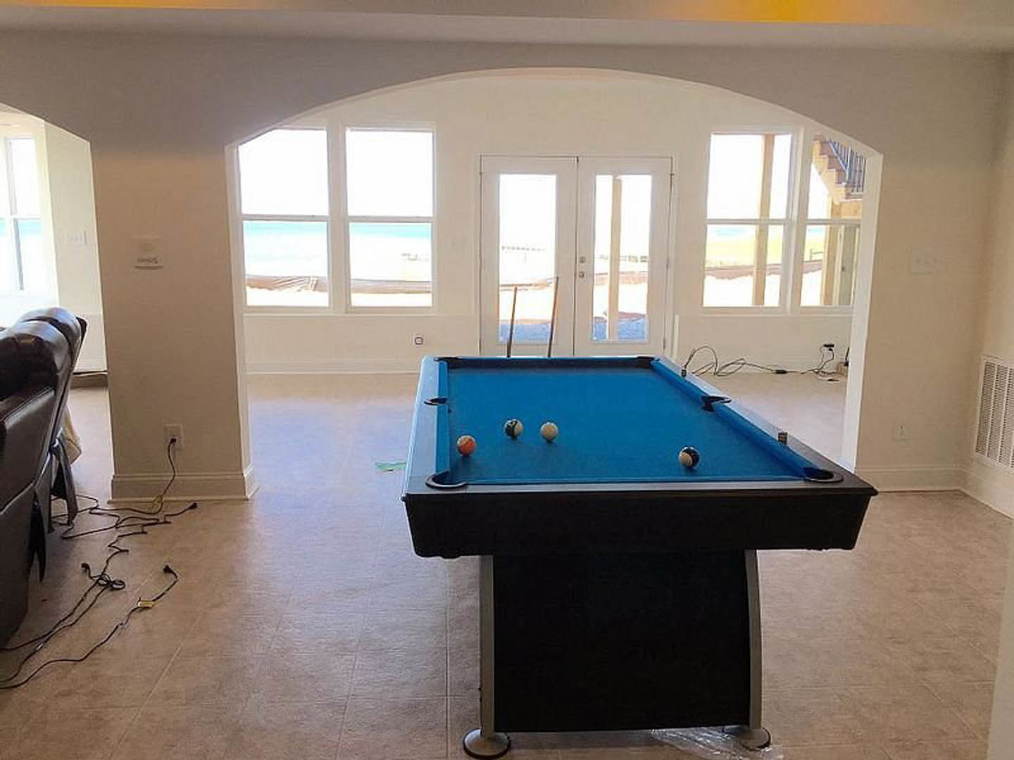 Pool table with billiard balls in a recreation room featuring double glass doors, light walls, and a chair nearby; black wires visible on the floor.