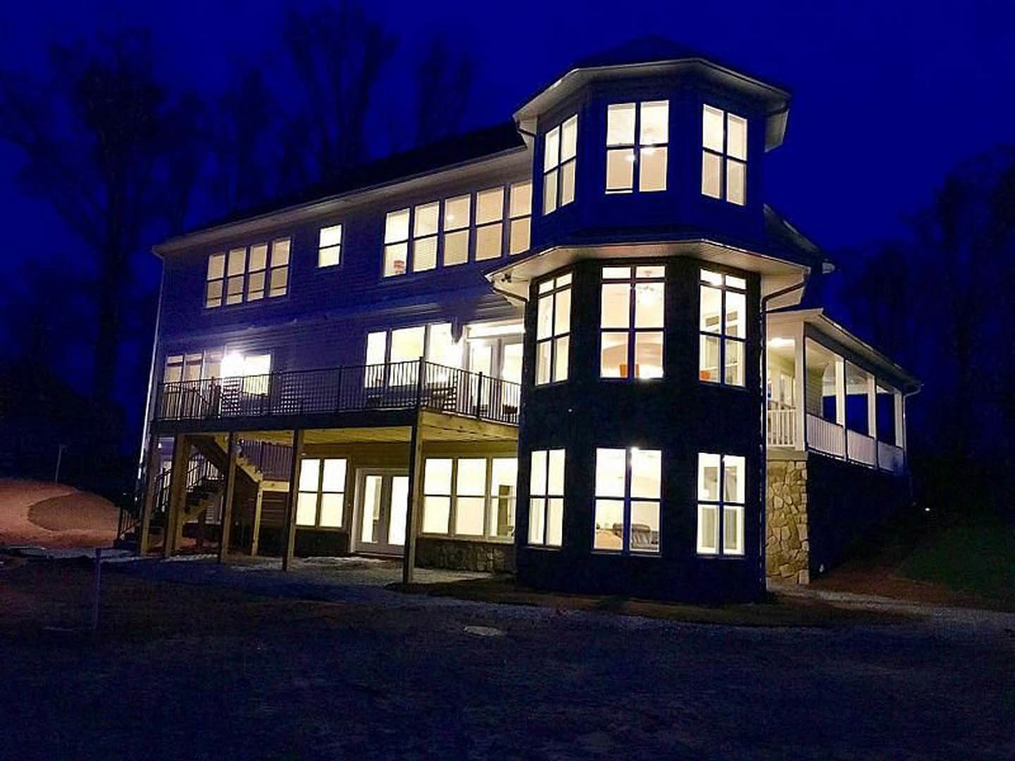 Two-story custom home with expansive wooden deck, covered front porch, multiple large windows, black metal railings, and exterior lighting at night.