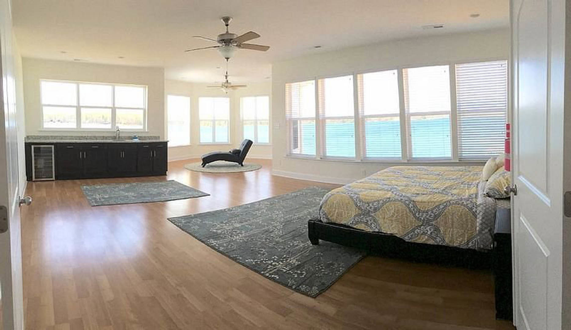 Spacious bedroom featuring a large bed with yellow and grey blanket, patterned rug on wood laminate flooring, ceiling fan with light fixture, window with white blinds, and