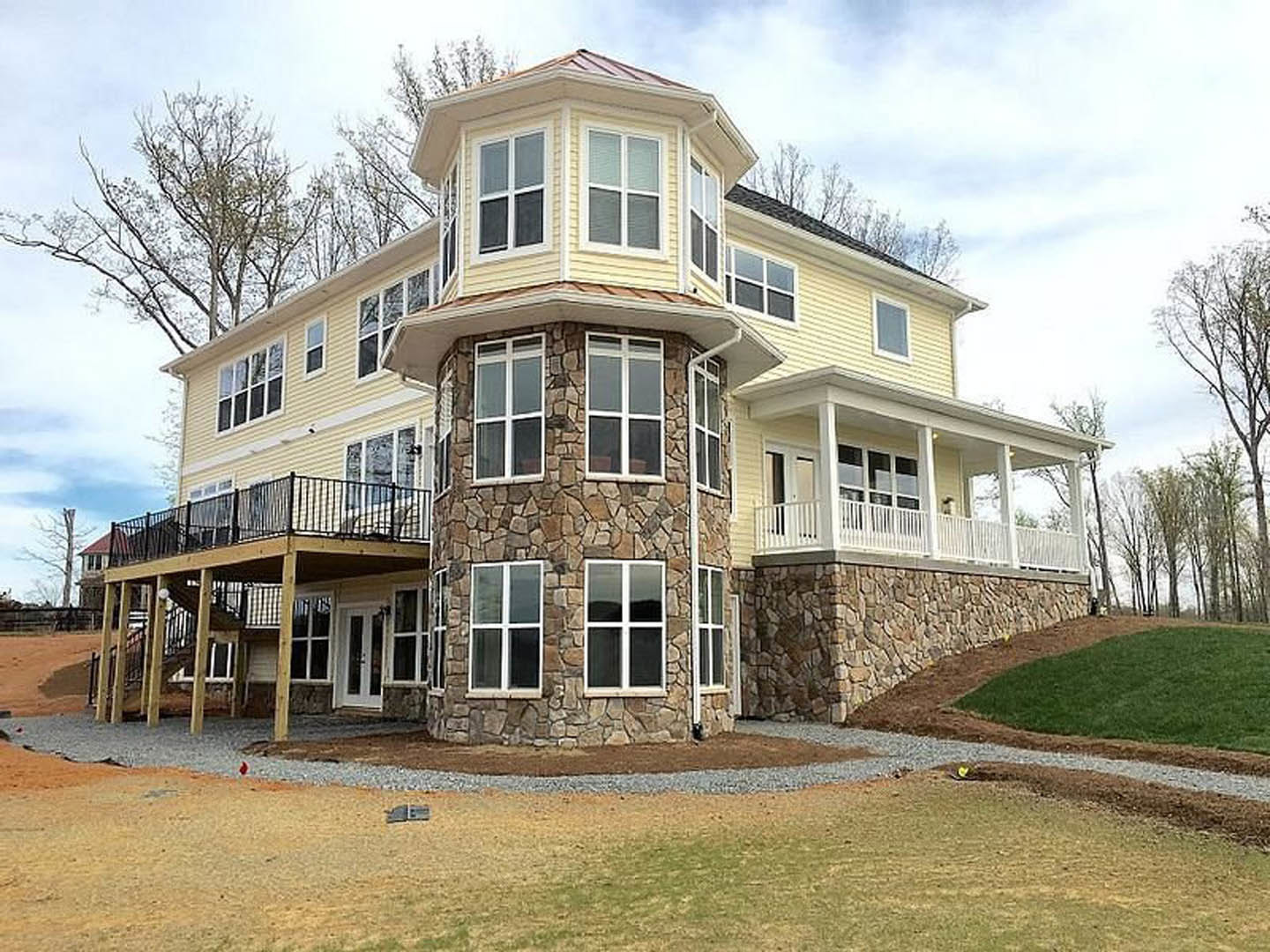 Expansive custom home with stone exterior walls, spacious stone patio, white-framed windows, upper balcony with white railing, manicured lawn in foreground, surrounded by trees
