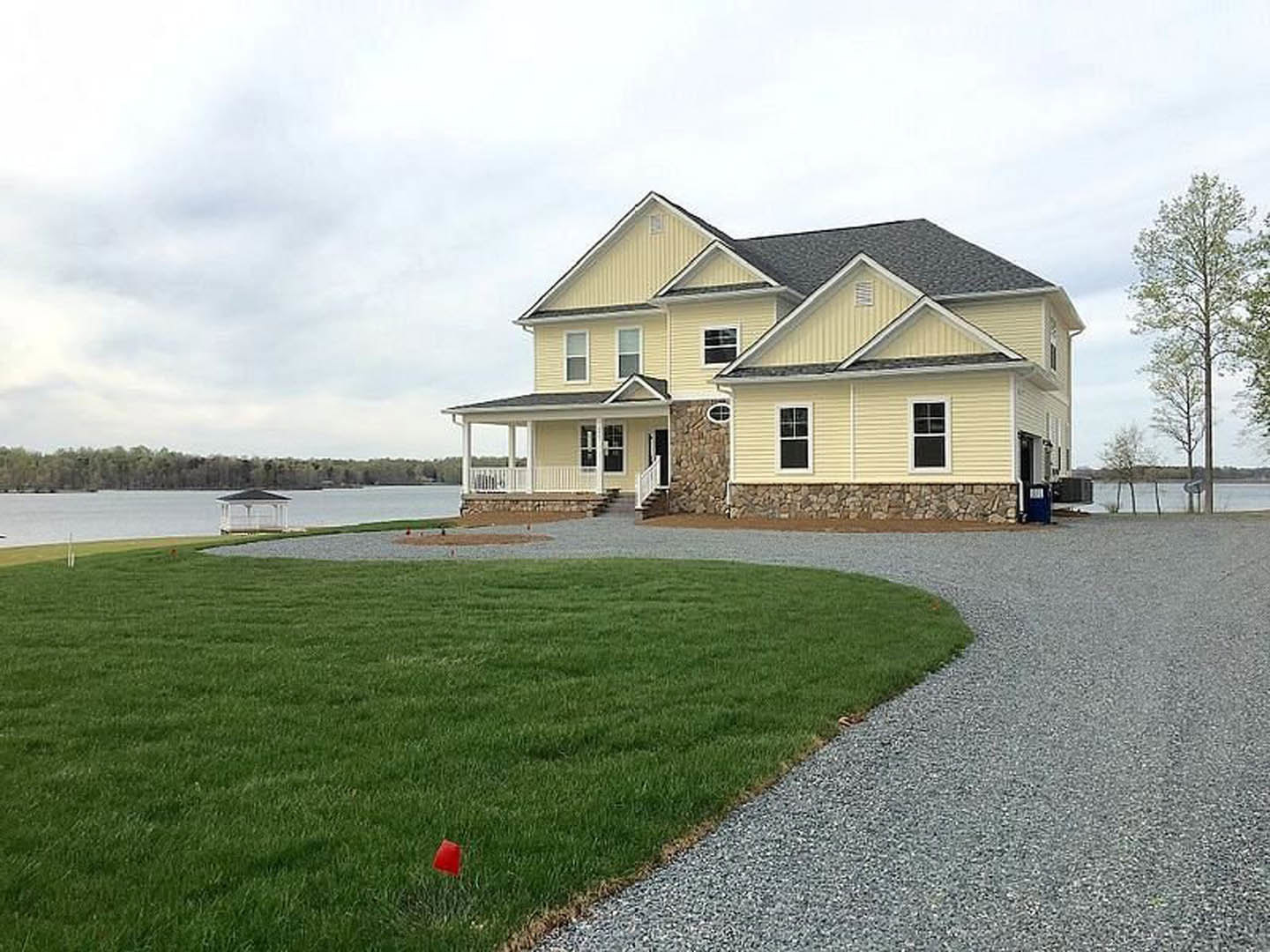 Yellow house with stone pillars, expansive green lawn, wide driveway, tall leafy tree, white building with black roof in background, red cone and red square on grass.