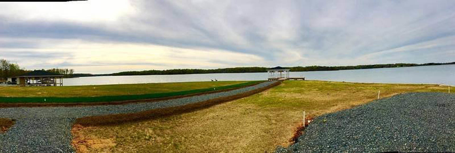 Grassy lawn leading to a wooden dock on a lake, surrounded by trees under a cloudy sky