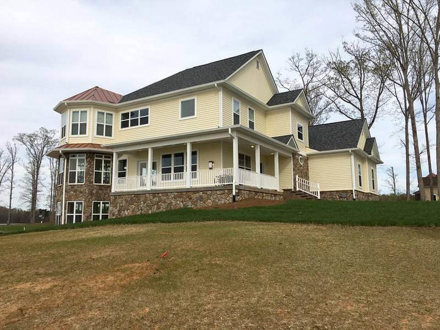 Yellow two-story house with stone exterior walls and porch, large windows, leafless trees in background, red object on grassy lawn