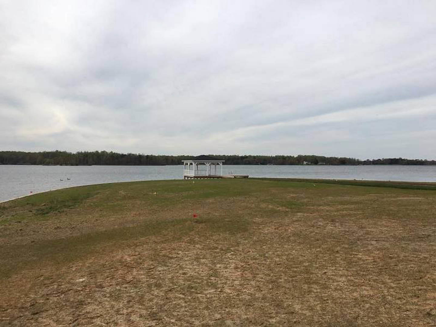 Grassy lawn bordered by a white building with black roof, white fence, and dock extending toward water under a cloudy sky; red object visible in grass.