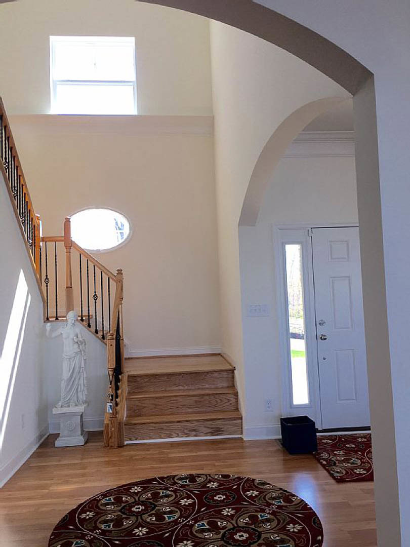 Wood staircase with white risers and handrail, patterned rug at base, statue of a woman in a white dress beside steps, black square object on hardwood floor, sunlight streaming