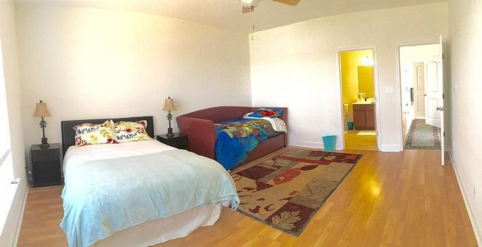 Bedroom with a bed featuring a white blanket, red chair beside it, couch draped with a colorful blanket, patterned rug on the floor, palm tree lamp, and a blurry blue bucket in the
