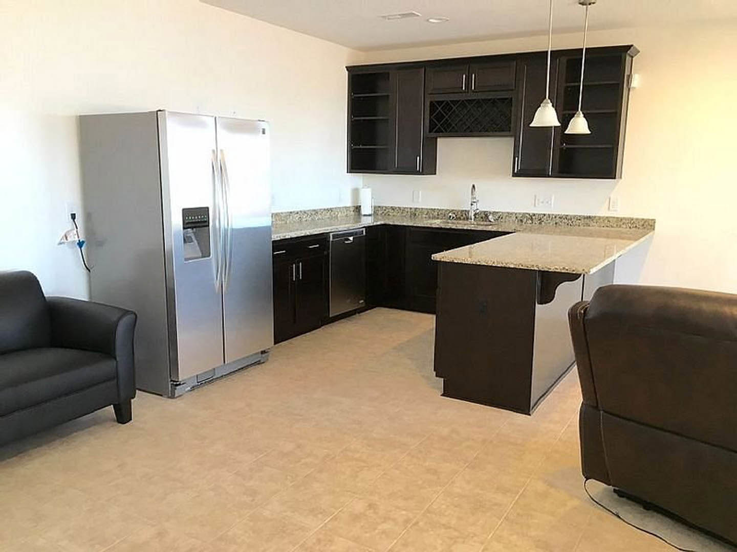 Spacious kitchen featuring a large central island with a leather chair, silver refrigerator, white cabinetry, wood flooring, and pendant lights; black couch visible in adjacent