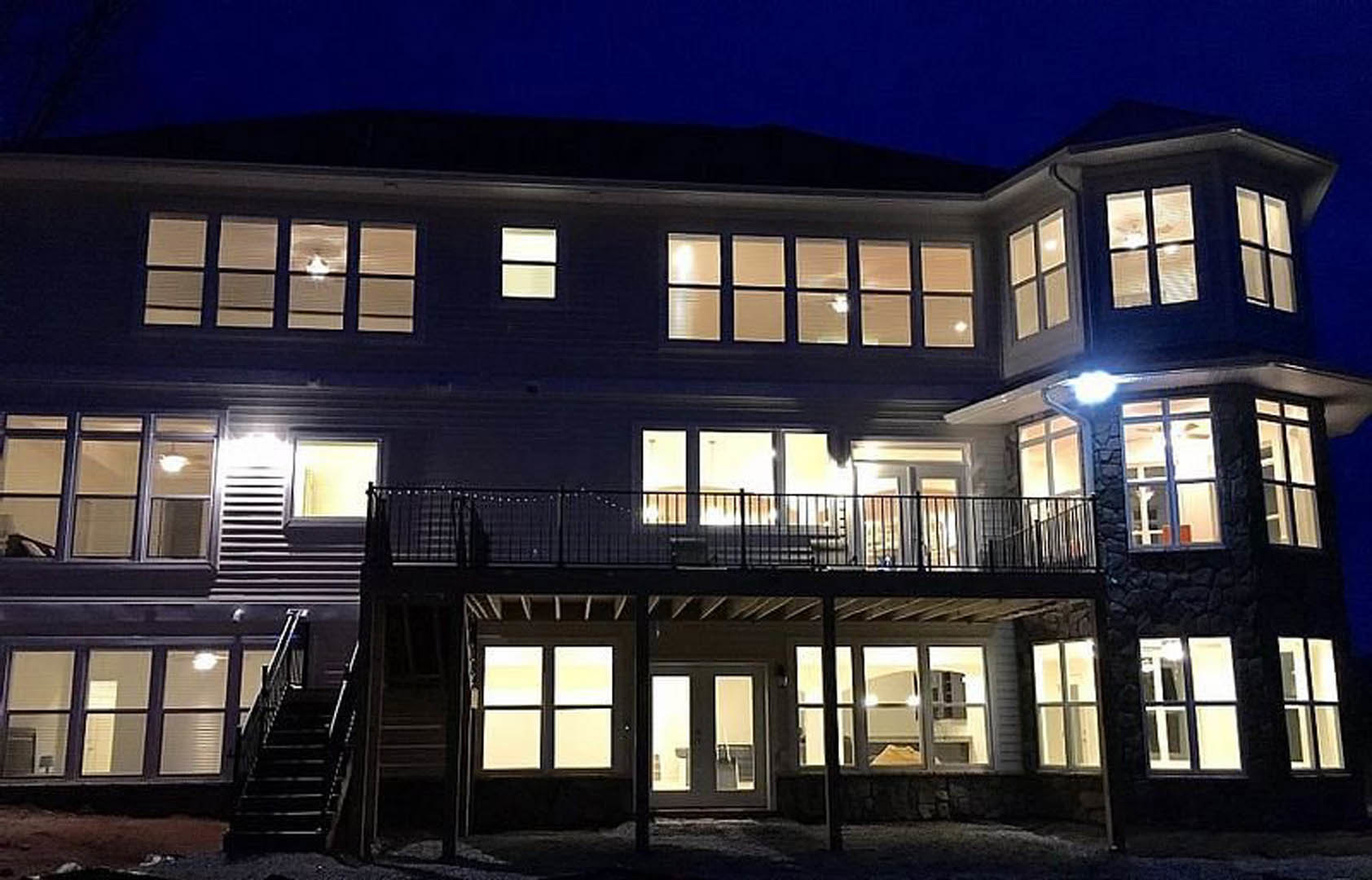 Modern two-story house with wood deck, metal railing, exterior stairs, large windows, and illuminated entry at dusk