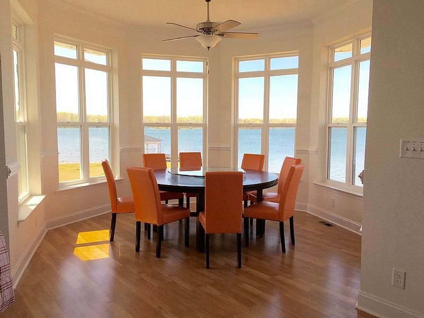 Dining room with wood flooring, rectangular table surrounded by upholstered chairs, ceiling fan with light fixture overhead, large window letting in natural light, neutral wall