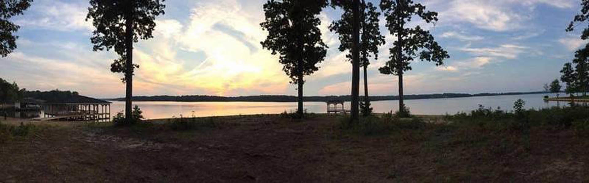 Sunset sky reflected on lake behind silhouetted tree, with bridge visible in the background and shoreline in foreground