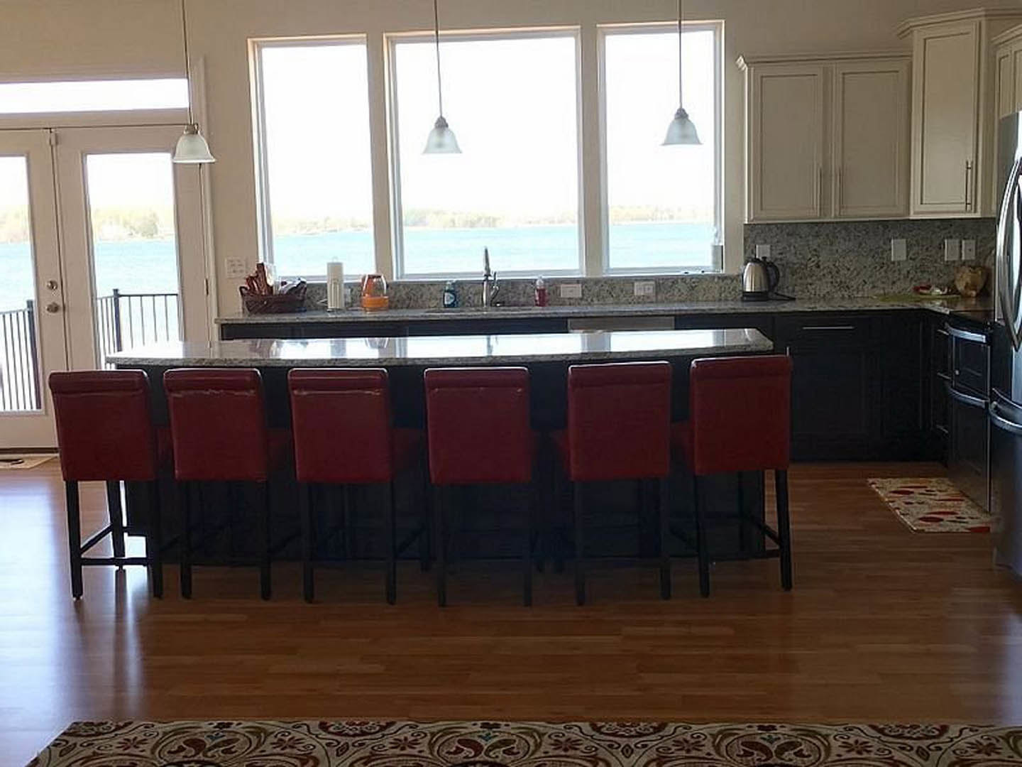 Open kitchen with light wood cabinetry, stone countertop bar, two modern chairs, large windows framing ocean view, silver kettle on counter, red chair with black legs, hardwood