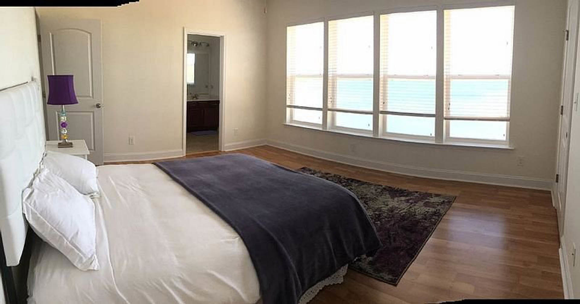 Spacious bedroom featuring a large bed with blue blanket and white pillows, patterned rug, purple lamp shade, row of windows with blinds, and partial view of bathroom with mirror