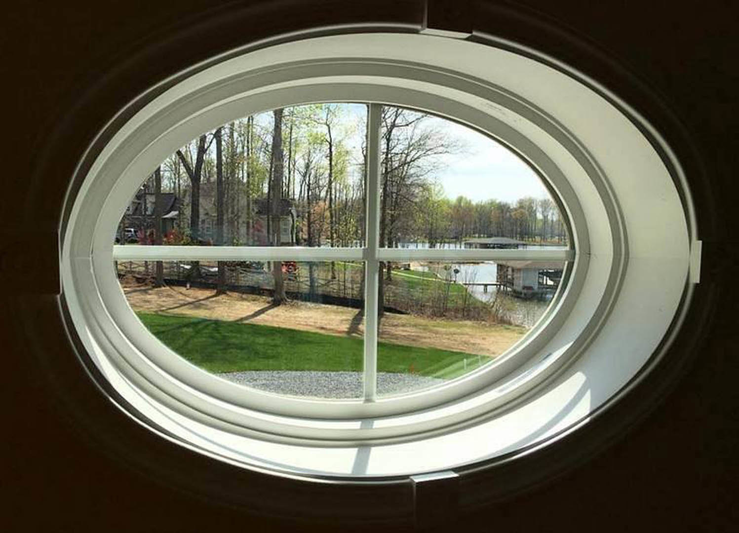 Round porthole window framed in white, overlooking a grassy field, lake, and trees in daylight