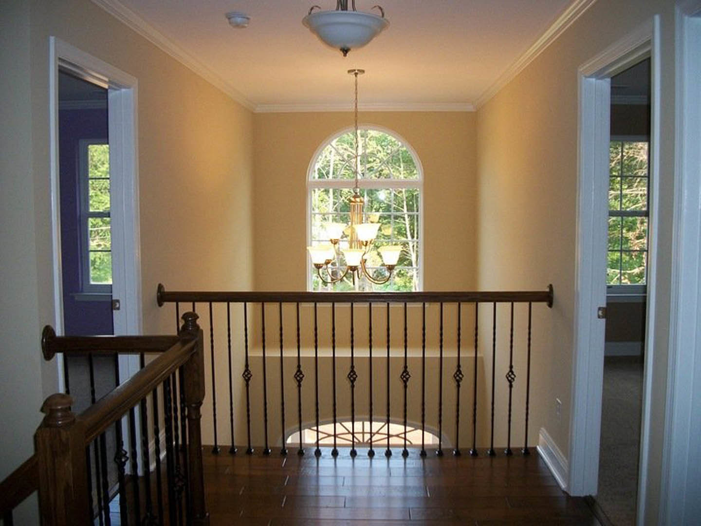 Wooden staircase with decorative railing, large window, and crystal chandelier hanging from ceiling; open doorway leads to hallway with natural light.