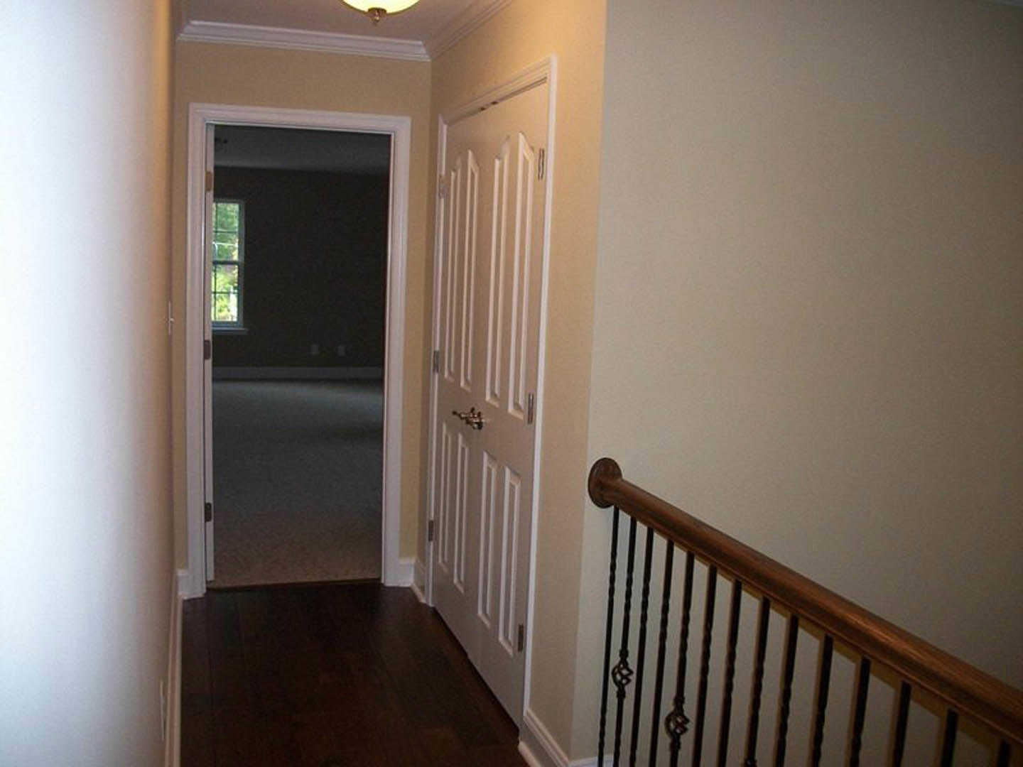 Hallway with dark wood flooring, white trim, white paneled doors, and a white-painted railing with balusters; window lets in natural light.