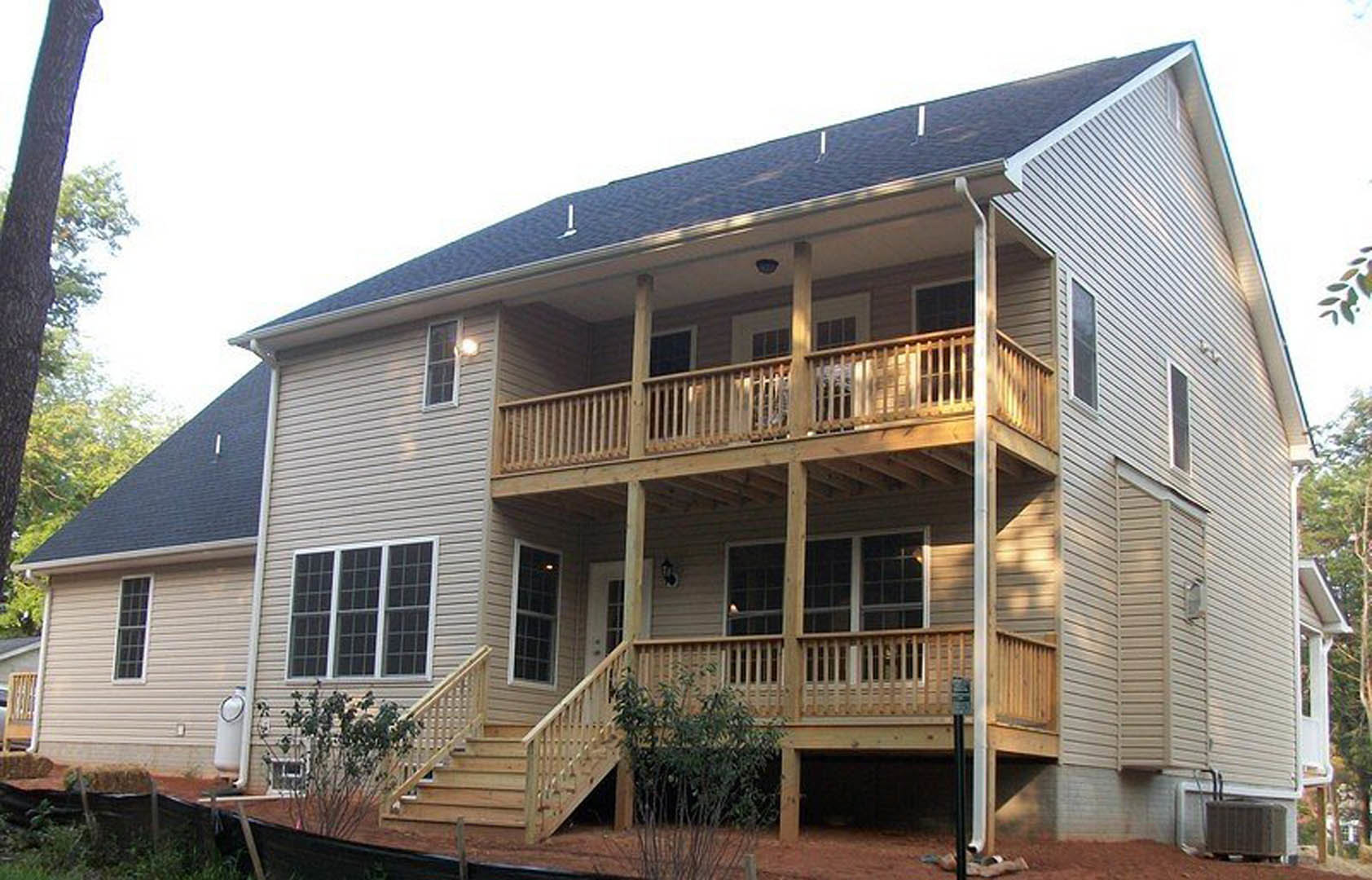 Two-story home with light siding, covered front porch, wooden deck with railing, stairs leading to landscaped yard, large windows, and potted plants near entry.