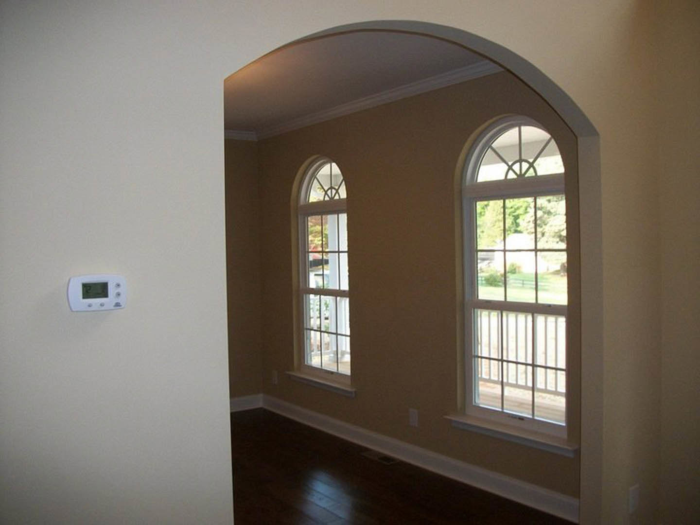 Arched windows with white trim, dark hardwood floor with floor vent, sunlight streaming onto wood planks, white electronic device mounted on wall, exterior view through window with