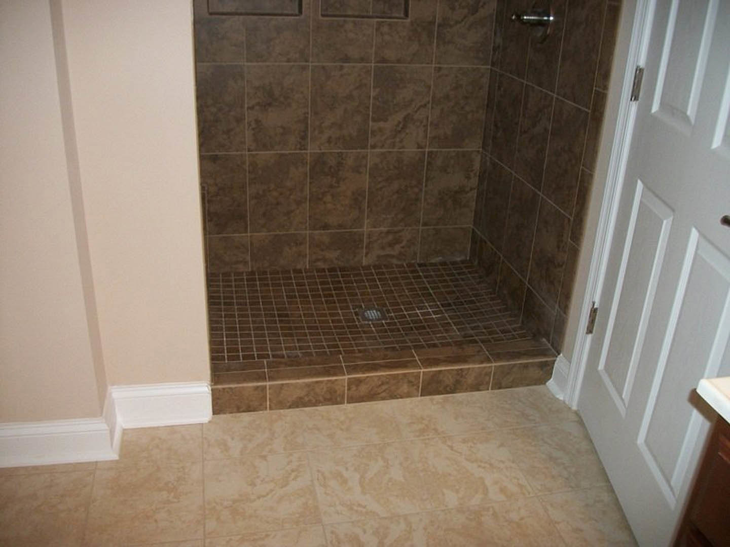 Brown tile shower with matching tile floor, metal drain, and partial view of glass shower door