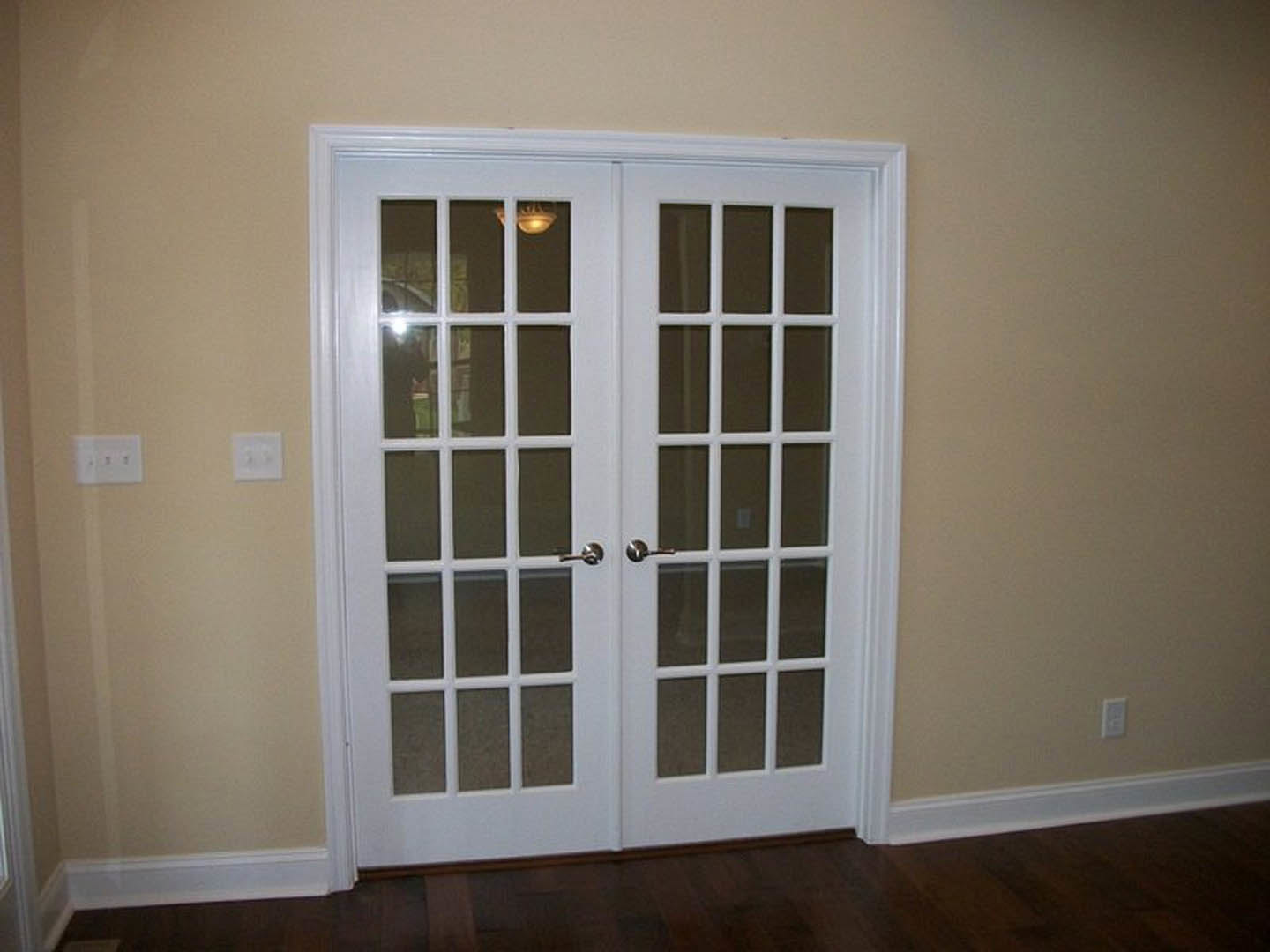 Double glass-paneled doors with dark wood flooring, white wall outlet with black switches, white light switch on a white wall, and contrasting black wall accents
