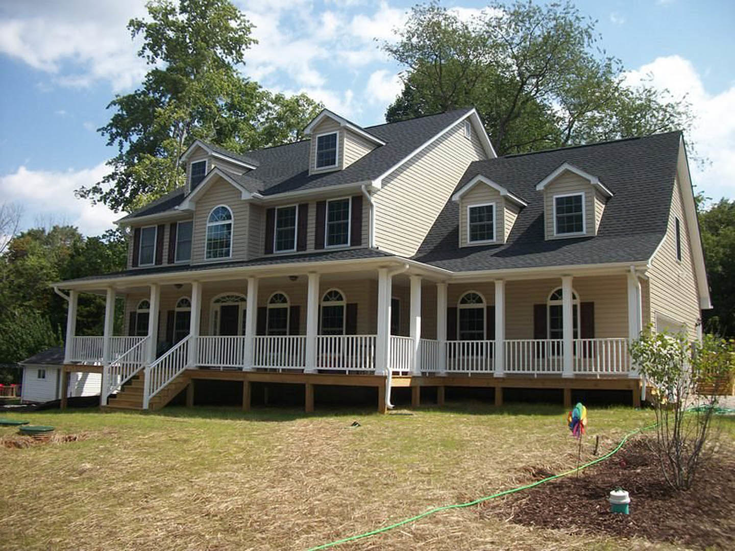 Expansive two-story home with wide covered porch, white columns, gray siding, multiple windows, manicured lawn, and mature tree in foreground