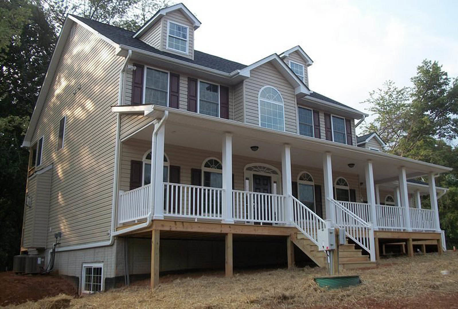 Two-story house with light-colored siding, covered front porch, white framed windows, wooden stairs, green lawn, and trees in the background