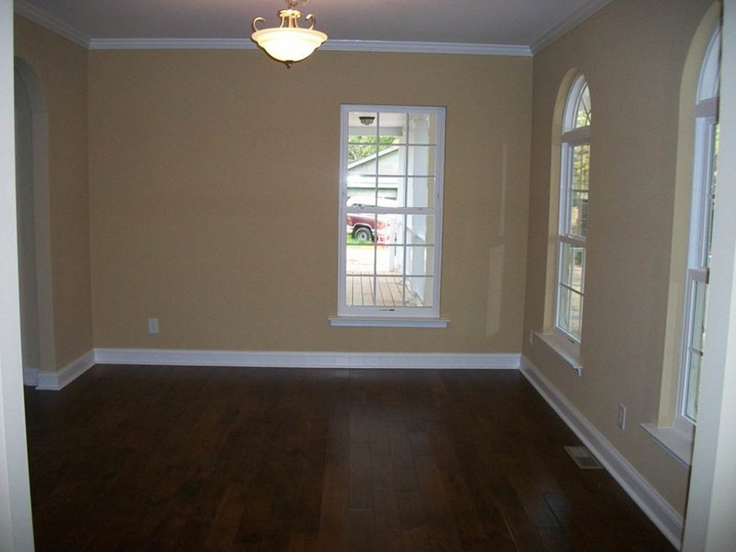 Room with dark hardwood floor, white baseboard trim, large window with view of parked car outside, white plaster walls, and ceiling light fixture with white shade