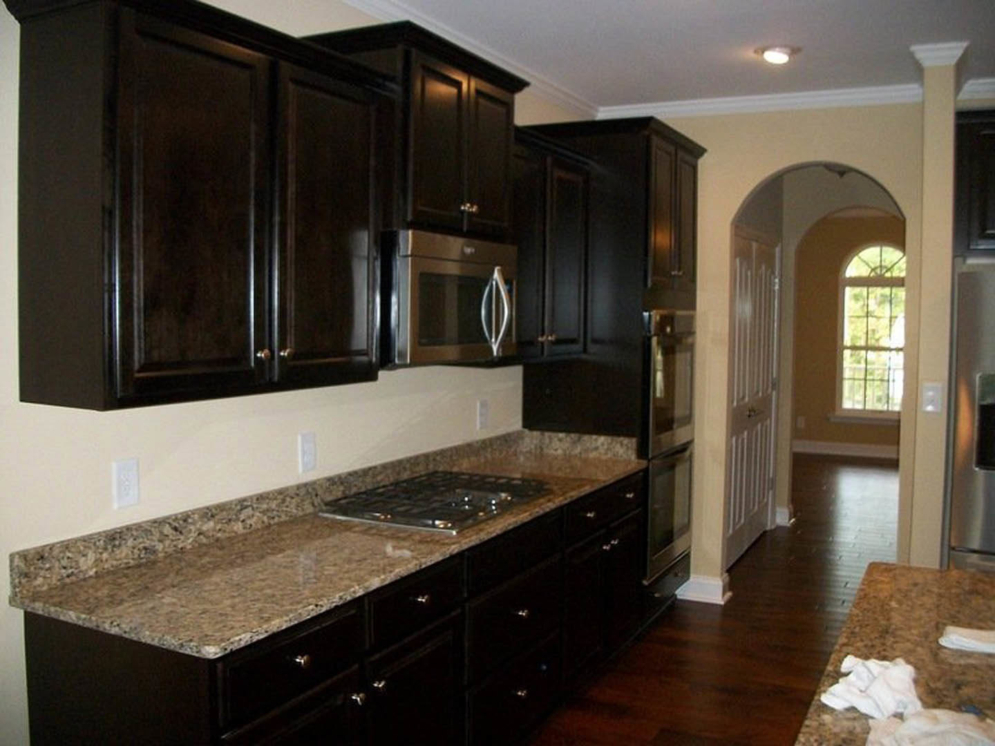 Dark wood kitchen cabinets, granite countertops, stainless steel microwave and stovetop, white outlet with cross detail, white towel on marble counter