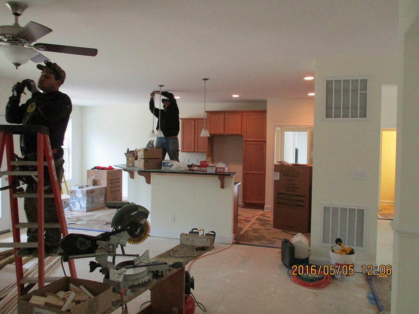 Two men installing a ceiling fan in a modern kitchen with white cabinetry, stainless steel appliances, a vent, a cardboard box, and a circular saw on the counter.