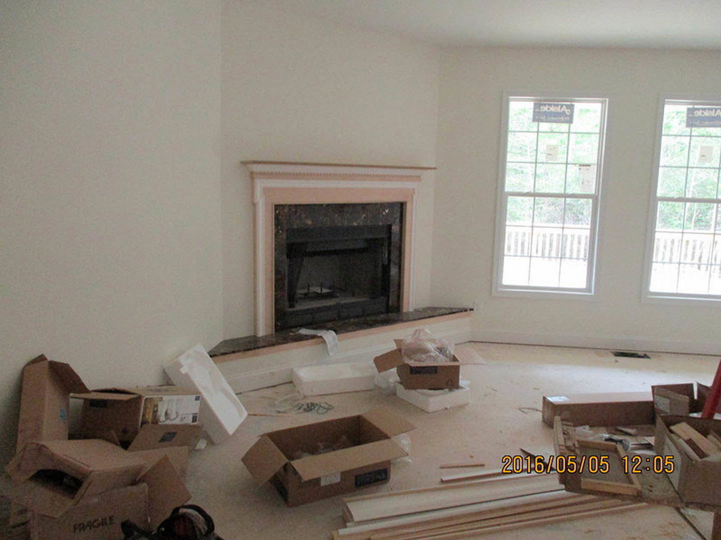 Living room with stone fireplace, white-framed windows displaying signs, cardboard boxes scattered on hardwood floor, digital scale atop one box