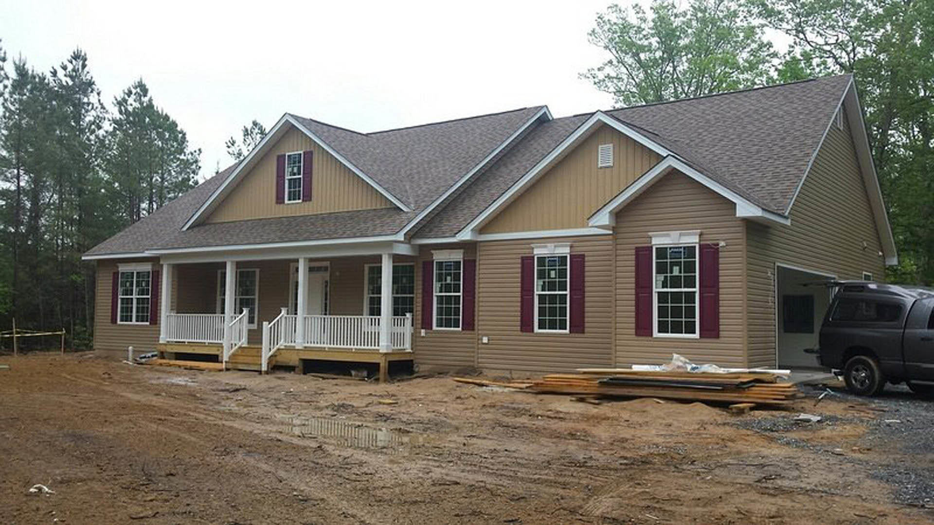 Partially built house with white porch railing, multi-pane windows, grey truck parked on dirt road, muddy ground with puddle, trees and sky in background
