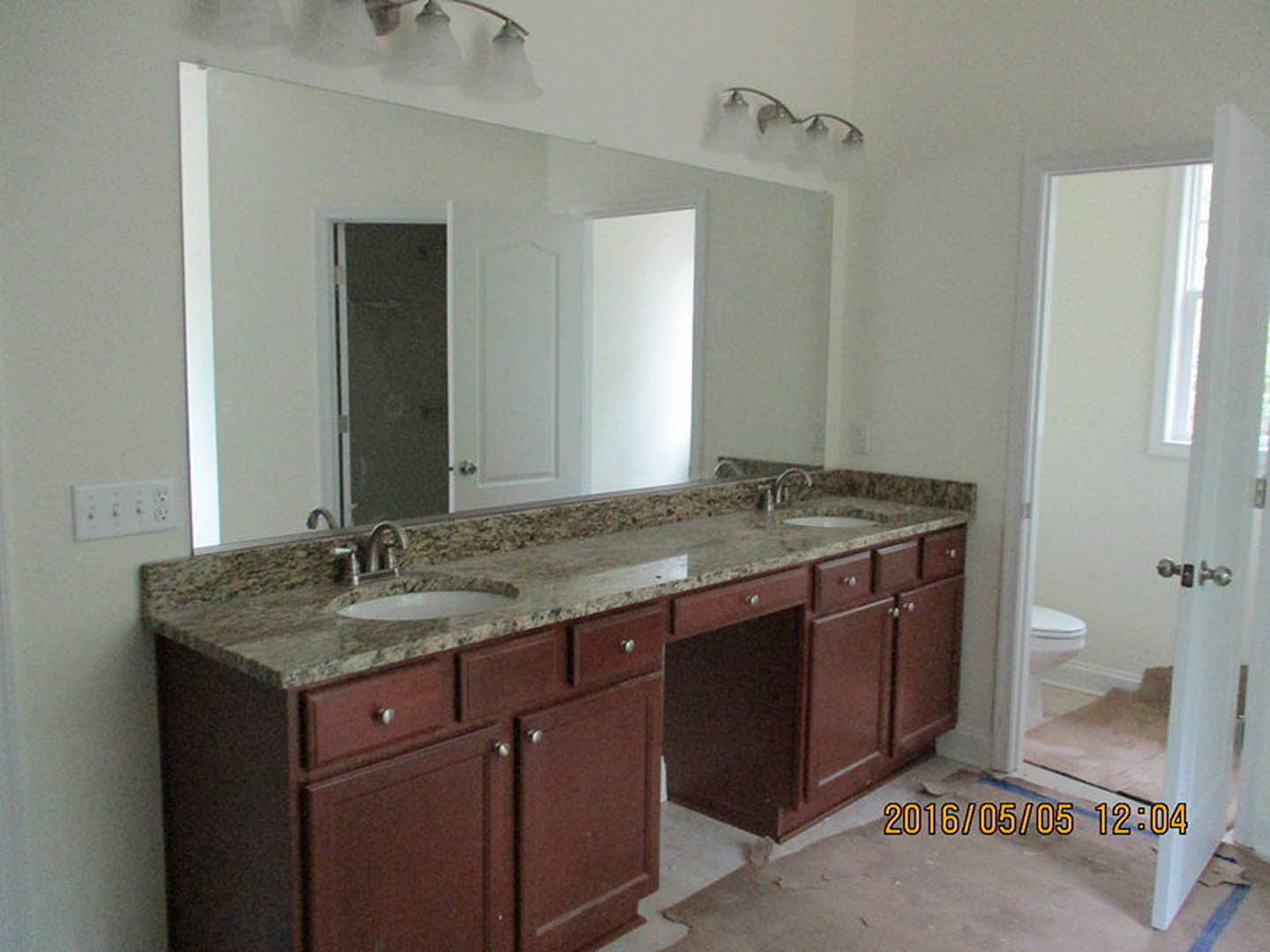 Bathroom with double sinks, marble countertop, white cabinetry, chrome faucets, tiled backsplash, white toilet, brown wooden door, and visible electrical outlet