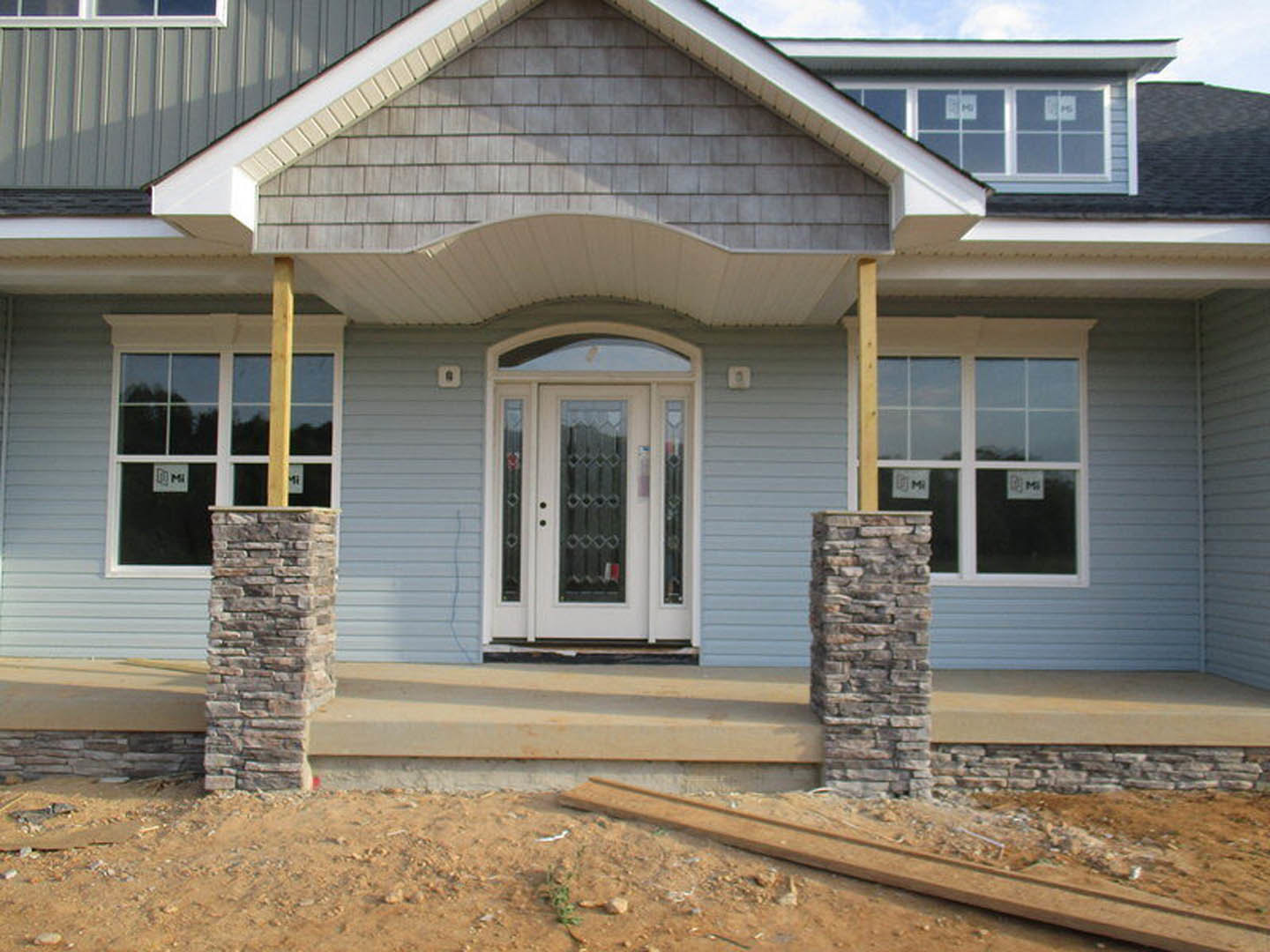 Stone pillar and white glass-paneled door on covered porch, gray siding exterior, sash windows, and unfinished construction elements visible
