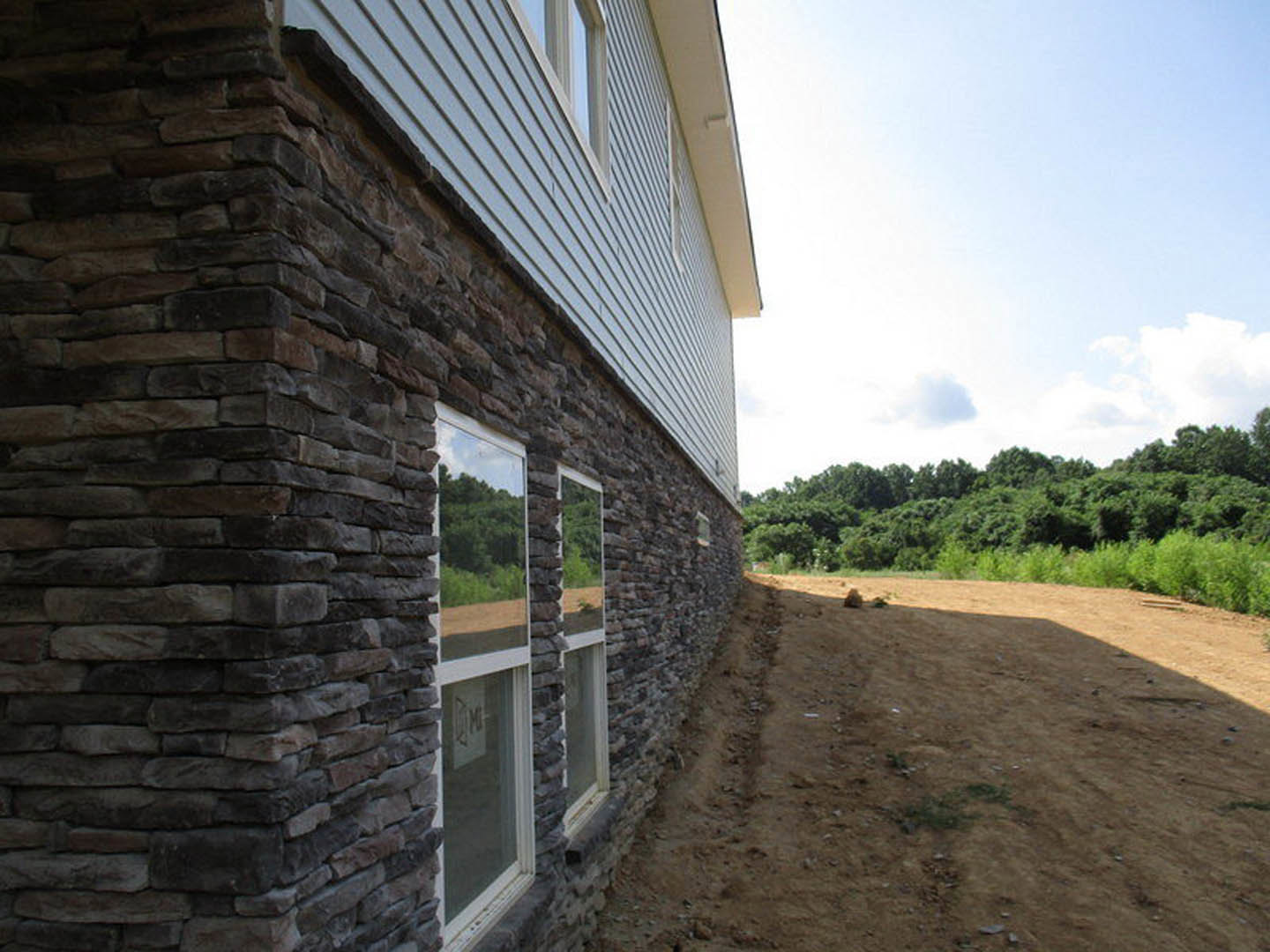 Side view of a brick exterior home with large windows, stone accents, and a wooden door, set beside a dirt field and surrounded by trees under a partly cloudy sky