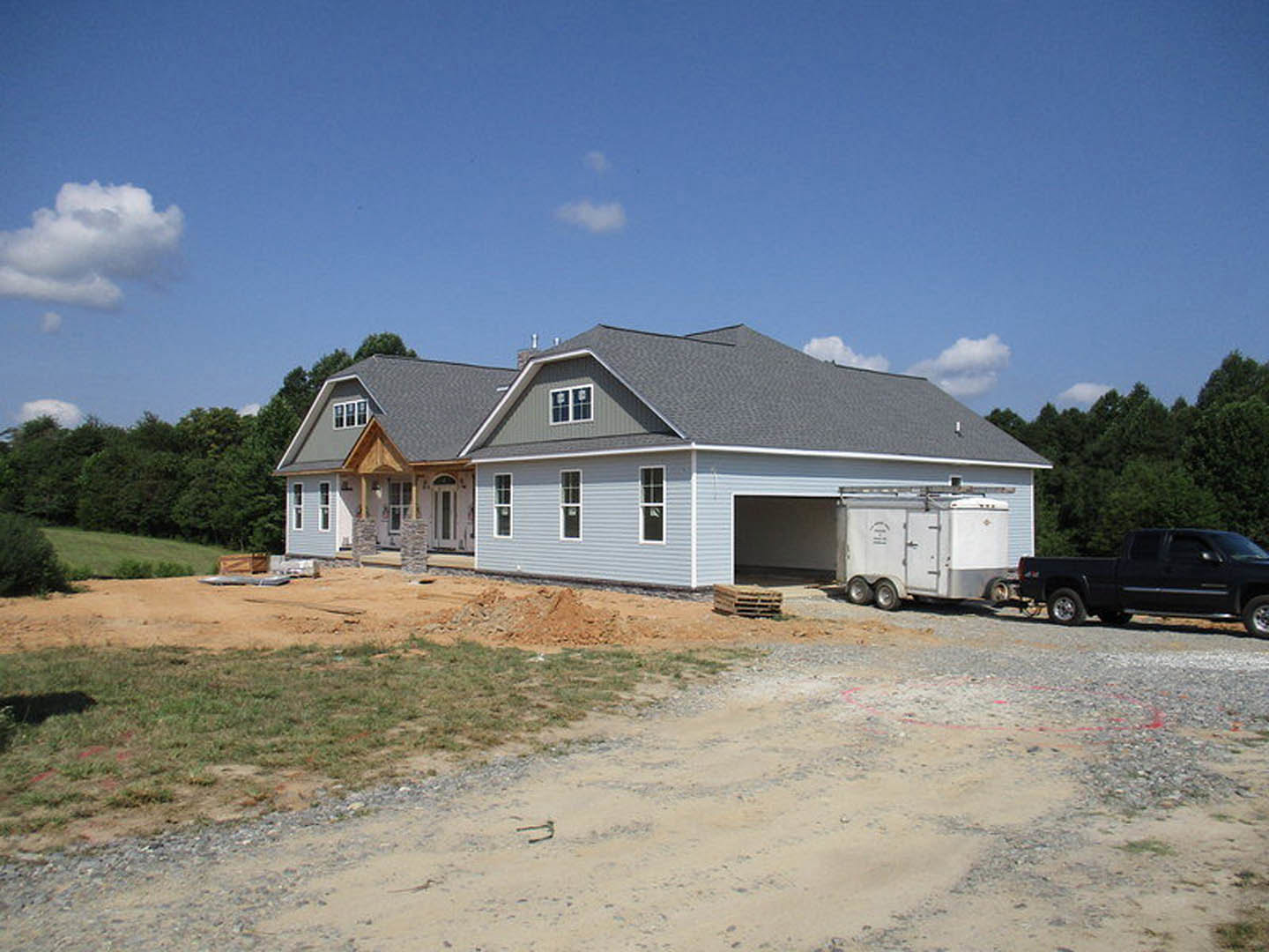 Framed house under construction with exposed plywood, black pickup truck parked on gravel driveway, attached garage, trailer nearby, window displaying permit sign, partly cloudy