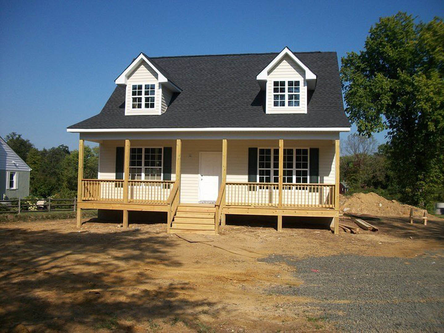 Partially built house with gray siding, white door, covered porch, dormer window, and leafy green tree in front