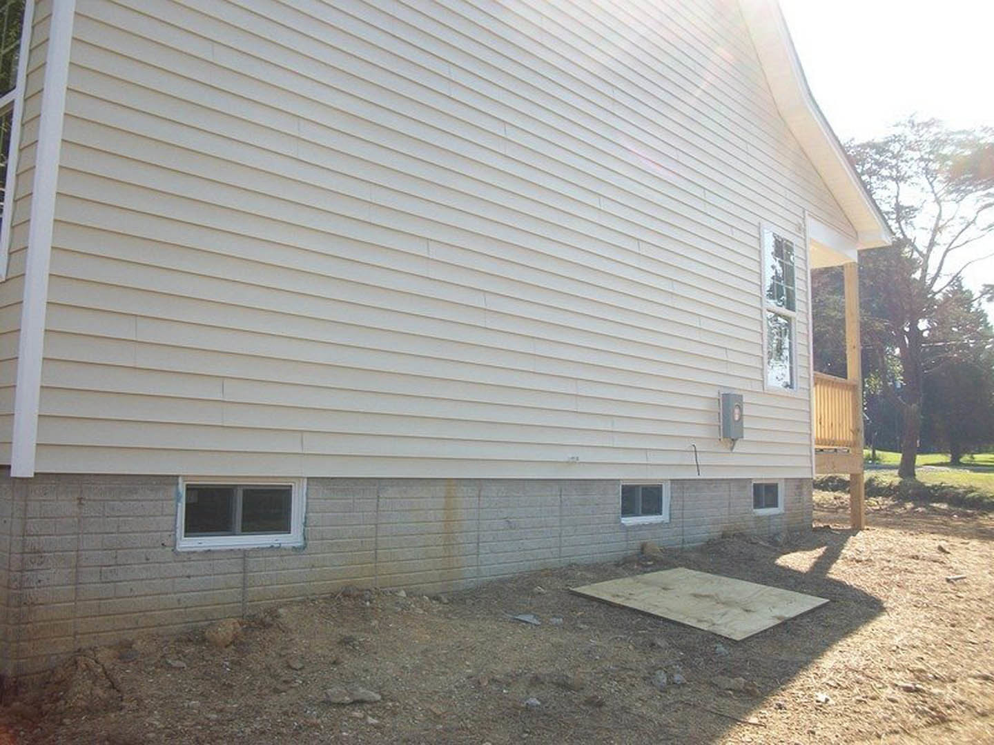 Front view of a house featuring a wooden deck and covered porch, white-framed windows, light-colored siding, grassy yard, and mature tree nearby.