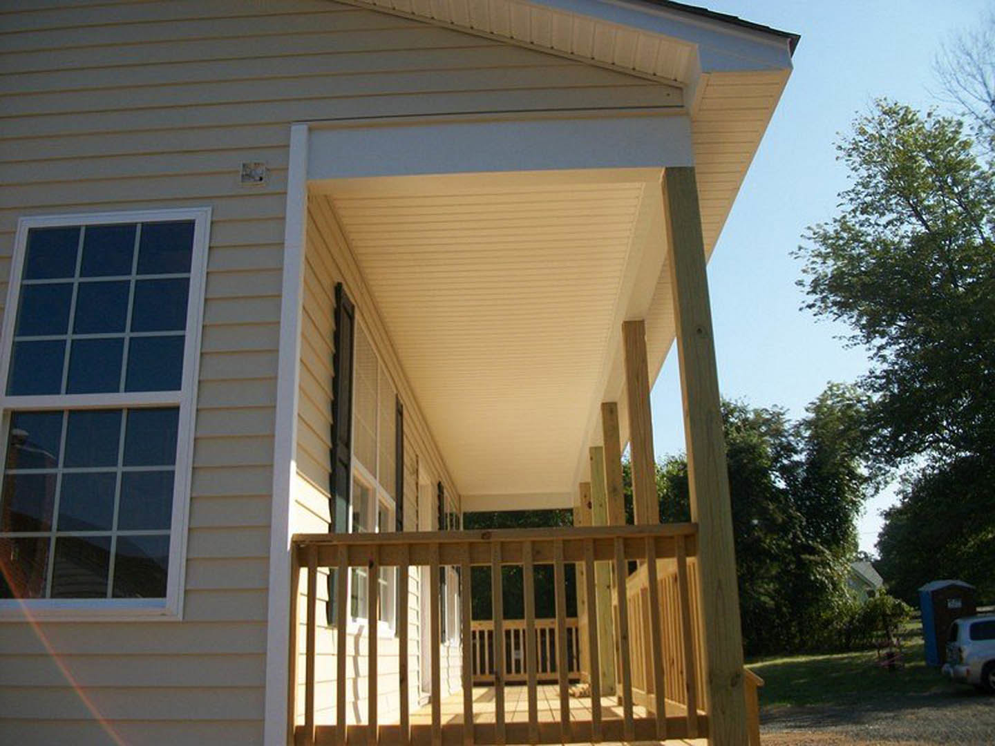 Covered porch with wooden railing, white siding, large window, shaded by nearby trees, partial view of a white car in the background