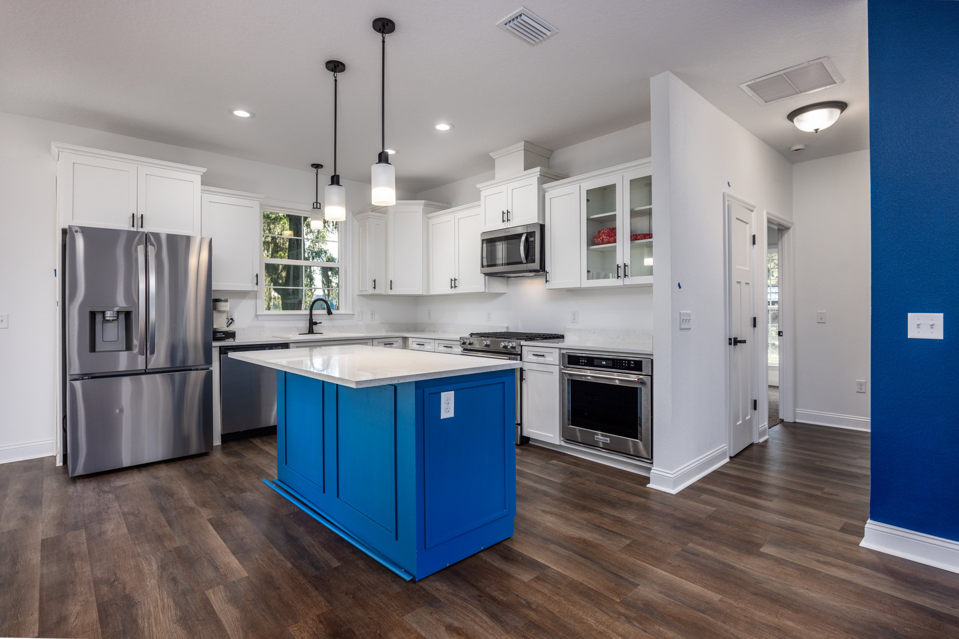 Blue kitchen island with white countertop, black faucet, stainless steel oven and microwave, refrigerator, white cabinetry, and light wood flooring.