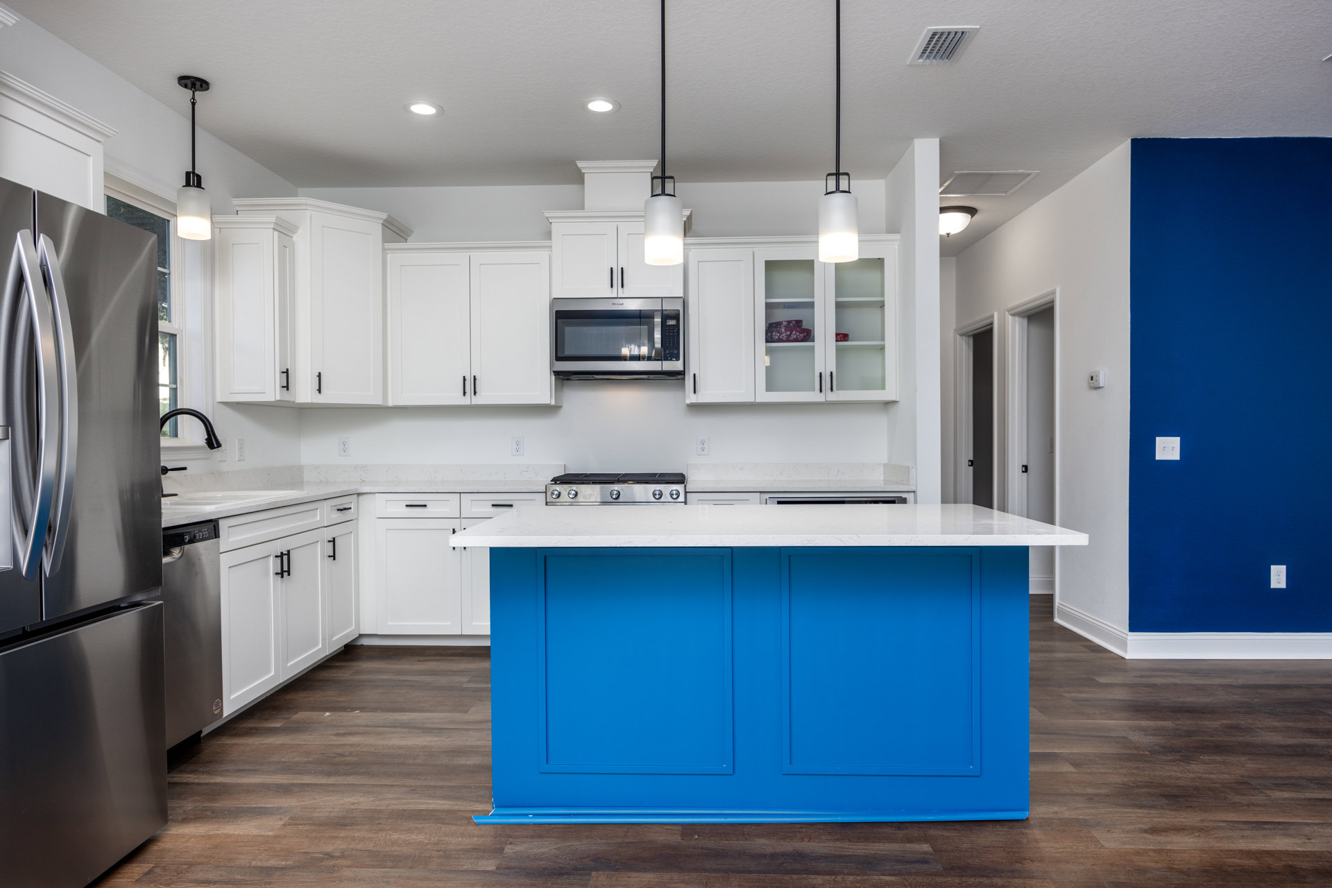 Blue kitchen island with white countertop, stainless steel appliances including refrigerator and microwave, white cabinetry, light fixture, and blue accent wall with white outlet.